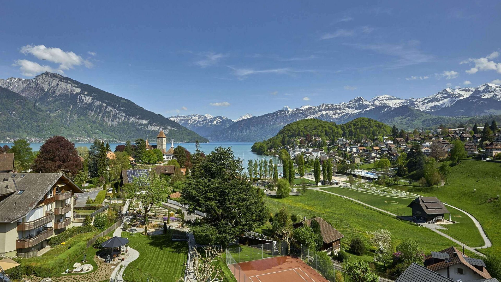 Aussicht auf den Garten und Tennisplatz des Hotels Eden sowie auf den Thunersee und die Bergkette dahinter