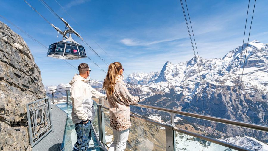 Eine Frau und ein Mann stehen auf der Thrill Walk Plattform und blicken zur Gondel vor Eiger, Mönch und Jungfrau