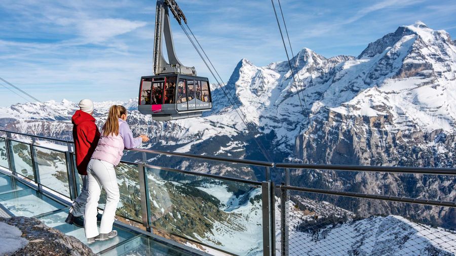Eine Frau und ein Mann stehen auf der Thrill Walk Plattform und blicken zur Gondel vor Eiger, Mönch und Jungfrau